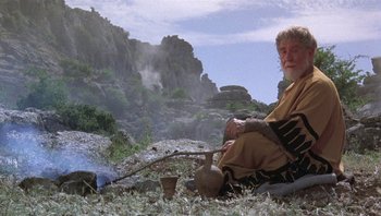 Movie still from “Clash of the Titans” (1981), directed by Desmond Davis – An older man sitting on the ground with a pot in his hand; Wide shot, High angle