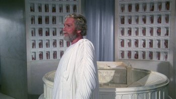 Movie still from “Clash of the Titans” (1981), directed by Desmond Davis – An older man wearing a white robe standing in front of a building; Medium shot, High angle