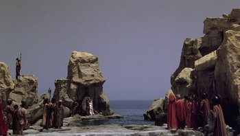Movie still from “Clash of the Titans” (1981), directed by Desmond Davis – A group of people standing on top of a beach near the ocean; Extreme Wide shot, High angle