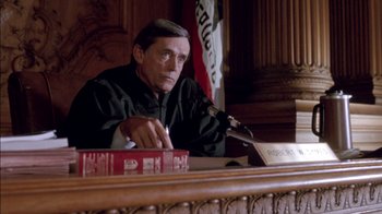 Movie still from “Class Action” (1991), directed by Michael Apted – A man sitting at a table in front of a microphone in a courtroom; Medium shot, High angle