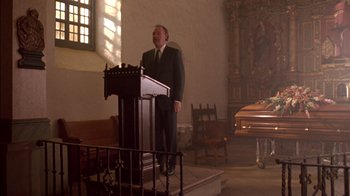 Movie still from “Class Action” (1991), directed by Michael Apted – A man in a suit standing at a podium in front of a casket; Wide shot, Low angle