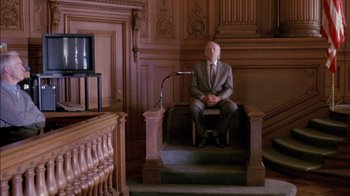Movie still from “Class Action” (1991), directed by Michael Apted – A man in a suit and tie sitting in a chair in front of a microphone; Wide shot, High angle