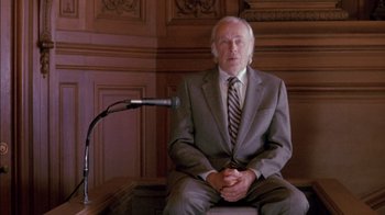 Movie still from “Class Action” (1991), directed by Michael Apted – An older man sitting in front of a microphone; Medium shot, Low angle