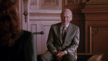 Movie still from “Class Action” (1991), directed by Michael Apted – An older man in a gray suit sitting in front of a microphone; Medium shot, Over the shoulder angle