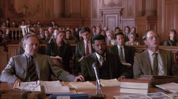 Movie still from “Class Action” (1991), directed by Michael Apted – A group of people sitting at a table in front of a microphone; Medium shot, High angle