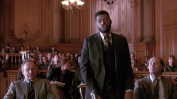 Movie still from “Class Action” (1991), directed by Michael Apted – A man standing in front of an audience wearing a suit and tie; Medium shot, Low angle