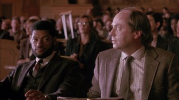 Movie still from “Class Action” (1991), directed by Michael Apted – A man in a suit and tie sitting in front of a crowd; Medium shot, Over the shoulder angle