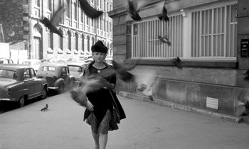 Movie still from “Cléo from 5 to 7” (1962), directed by Agnès Varda – A woman in a black dress is walking down the street with pigeons flying around her; Wide shot, Low angle