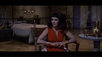 Movie still from “Cleopatra” (1963), directed by Rouben Mamoulian – A woman sitting in a chair wearing a red dress; Medium shot, Over the shoulder angle