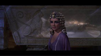 Movie still from “Cleopatra” (1963), directed by Rouben Mamoulian – A woman in a purple dress with a bunch of hair on her head; Close Up shot, Over the shoulder angle