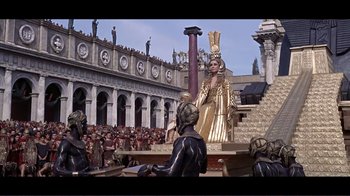 Movie still from “Cleopatra” (1963), directed by Rouben Mamoulian – A statue of a woman in a gold dress and a crowd of people in the background; Extreme Wide shot, High angle
