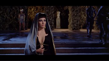 Movie still from “Cleopatra” (1963), directed by Rouben Mamoulian – A woman dressed as cleopatra standing in front of trees; Medium shot, Over the shoulder angle