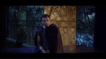 Movie still from “Cleopatra” (1963), directed by Rouben Mamoulian – A man wearing a black cape sitting in front of a wall; Medium shot, Over the shoulder angle
