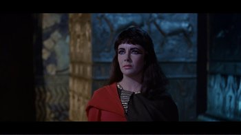 Movie still from “Cleopatra” (1963), directed by Rouben Mamoulian – A woman in a red and black outfit standing in front of a wall; Close Up shot, Over the shoulder angle