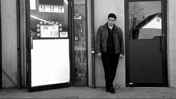 Movie still from “Clerks” (1994), directed by Kevin Smith – A black and white photo of a man standing in front of a store; Wide shot, Low angle