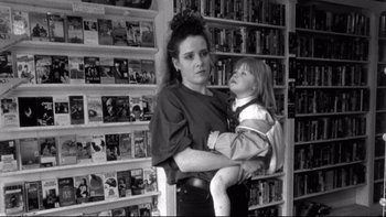 Movie still from “Clerks” (1994), directed by Kevin Smith – A woman holding a small child in her arms in front of a wall full of dvd's; Medium shot, High angle