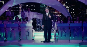 Movie still from “Click” (2006), directed by Frank Coraci – A man in a suit and tie on a stage; Wide shot, High angle