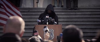Movie still from “Death Note” (2017), directed by Adam Wingard – A man in a black hooded jacket is speaking at a podium; Medium shot, Over the shoulder angle