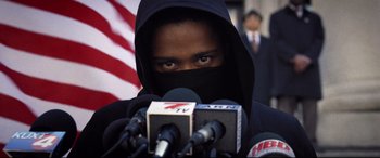Movie still from “Death Note” (2017), directed by Adam Wingard – A man wearing a black mask and covering his face while standing in front of microphones; Close Up shot, Low angle