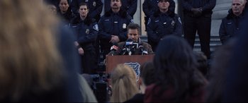 Movie still from “Death Note” (2017), directed by Adam Wingard – A group of police officers standing behind a man speaking at a press conference; Medium shot, Over the shoulder angle