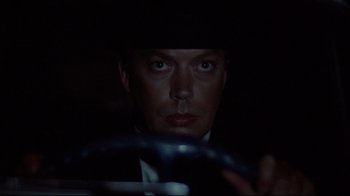 Movie still from “Clue” (1985), directed by Jonathan Lynn – A person in a suit driving a car; Close Up shot, Low angle