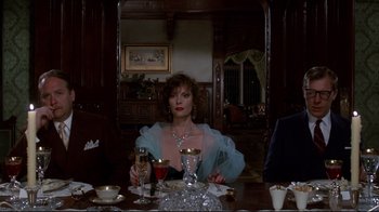 Movie still from “Clue” (1985), directed by Jonathan Lynn – A woman sitting at a dinner table with a glass of wine in front of her; Medium shot, Low angle