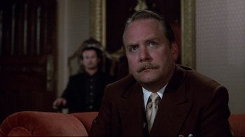 Movie still from “Clue” (1985), directed by Jonathan Lynn – A man in a suit and tie sitting on a couch; Close Up shot, Low angle
