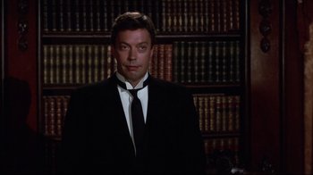 Movie still from “Clue” (1985), directed by Jonathan Lynn – A man in a suit and tie in front of a book shelf; Medium shot, Low angle