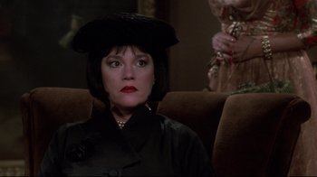 Movie still from “Clue” (1985), directed by Jonathan Lynn – A woman in a black hat sitting on a brown couch; Close Up shot, Over the shoulder angle