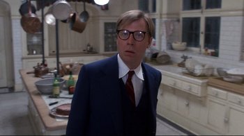 Movie still from “Clue” (1985), directed by Jonathan Lynn – A man wearing a suit and tie in a kitchen; Medium shot, Over the shoulder angle