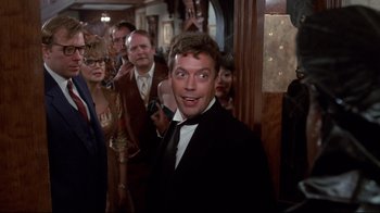 Movie still from “Clue” (1985), directed by Jonathan Lynn – A man in a tuxedo standing in front of a group of people; Medium shot, Over the shoulder angle