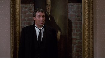 Movie still from “Clue” (1985), directed by Jonathan Lynn – A man wearing a suit and tie standing in front of a painting; Medium shot, Over the shoulder angle