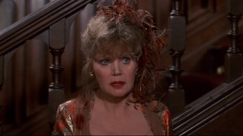 Movie still from “Clue” (1985), directed by Jonathan Lynn – An older woman wearing a red and gold dress and a feather headpiece; Close Up shot, Over the shoulder angle