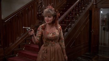 Movie still from “Clue” (1985), directed by Jonathan Lynn – A woman in a dress holding a gun in front of a staircase; Medium shot, High angle