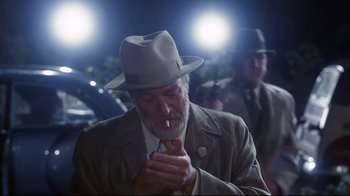 Movie still from “Clue” (1985), directed by Jonathan Lynn – An older man wearing a hat and smoking a cigarette; Medium shot, Over the shoulder angle
