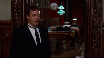 Movie still from “Clue” (1985), directed by Jonathan Lynn – A man in a suit standing in front of a man laying on a bed; Medium shot, Over the shoulder angle