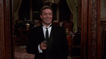 Movie still from “Clue” (1985), directed by Jonathan Lynn – A man in a suit and tie holding a camera; Medium shot, Over the shoulder angle