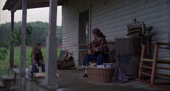 Movie still from “Coal Miner's Daughter” (1980), directed by Michael Apted – A woman sitting on a porch playing a guitar; Wide shot, Over the shoulder angle