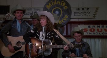 Movie still from “Coal Miner's Daughter” (1980), directed by Michael Apted – A woman in a cowboy hat playing a guitar in front of a microphone; Medium shot, Over the shoulder angle
