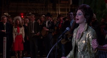 Movie still from “Coal Miner's Daughter” (1980), directed by Michael Apted – A woman in a gold dress singing into a microphone in front of a group of people; Medium shot, Over the shoulder angle
