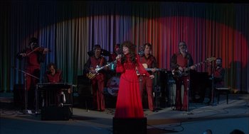 Movie still from “Coal Miner's Daughter” (1980), directed by Michael Apted – A woman in a long red dress singing into a microphone; Wide shot, High angle