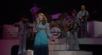 Movie still from “Coal Miner's Daughter” (1980), directed by Michael Apted – A woman singing into a microphone in front of a group of men; Wide shot, Low angle