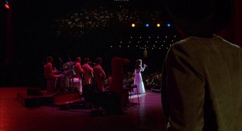Movie still from “Coal Miner's Daughter” (1980), directed by Michael Apted – A group of people on a stage with a woman singing into a microphone; Wide shot, Over the shoulder angle