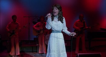 Movie still from “Coal Miner's Daughter” (1980), directed by Michael Apted – A woman in a long white dress singing into a microphone; Medium shot, Low angle