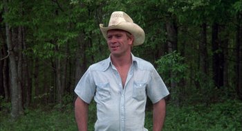 Movie still from “Coal Miner's Daughter” (1980), directed by Michael Apted – A man wearing a cowboy hat standing in front of a forest; Medium shot, Low angle