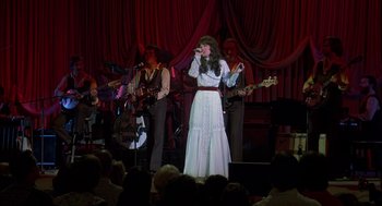 Movie still from “Coal Miner's Daughter” (1980), directed by Michael Apted – A woman in a long white dress singing into a microphone; Wide shot, Low angle
