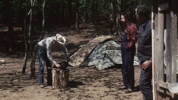 Movie still from “Cockfighter” (1974), directed by Monte Hellman – A man chopping wood in the woods while another man looks on; Wide shot, High angle