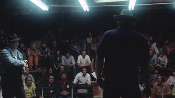 Movie still from “Cockfighter” (1974), directed by Monte Hellman – A crowd of people sitting in a dark room; Wide shot, Over the shoulder angle
