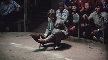 Movie still from “Cockfighter” (1974), directed by Monte Hellman – A man kneeling down to pet a rooster; Wide shot, High angle