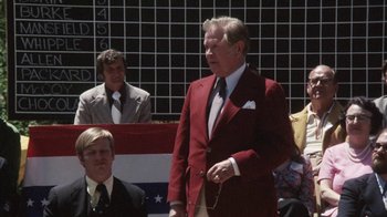 Movie still from “Cockfighter” (1974), directed by Monte Hellman – An older man in a red suit stands in front of an american flag; Medium shot, Over the shoulder angle